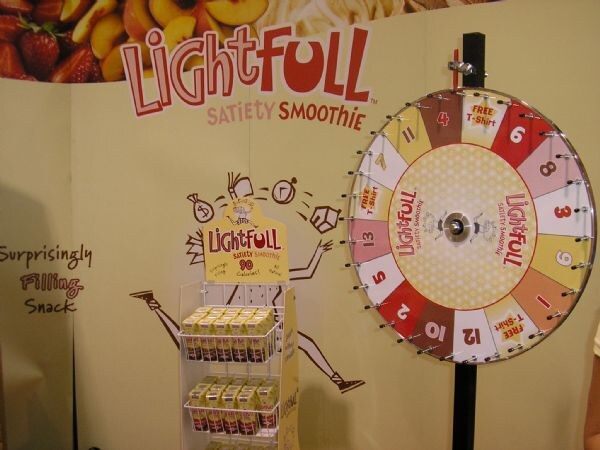 A Lightfull Satiety Smoothie booth with a prize wheel numbered 1 to 13 with Free T-Shirt spaces, plus a rack of smoothie cartons in front of a wall graphic.
