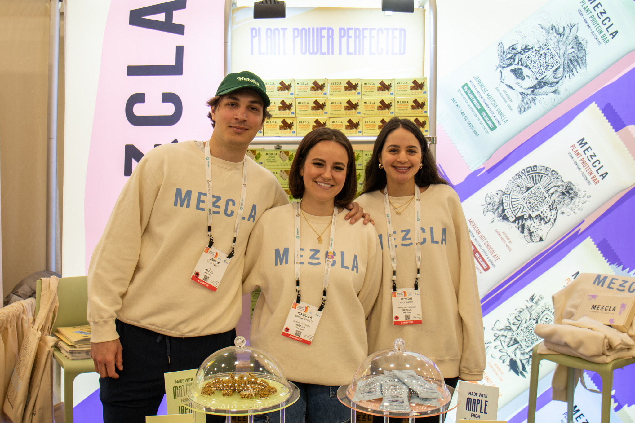 Three smiling people in tan Mezcla sweatshirts stand in front of Mezcla plant-based protein bar displays