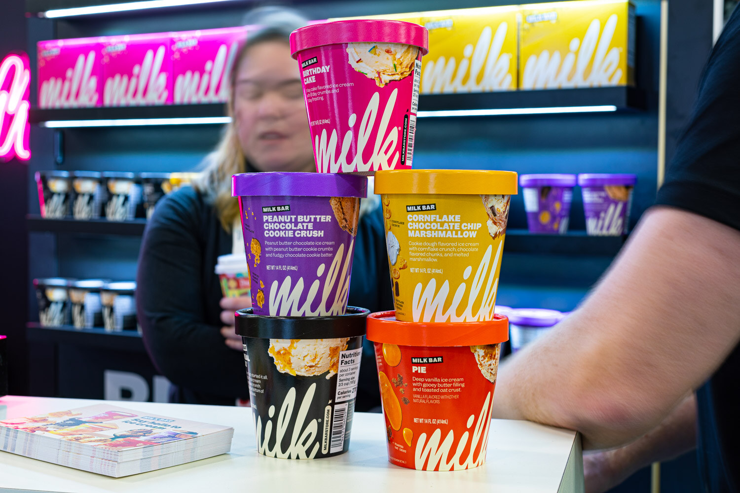 Pints of Milk Bar ice cream in pink, purple, yellow and orange are stacked on a counter in front of a neon milk sign