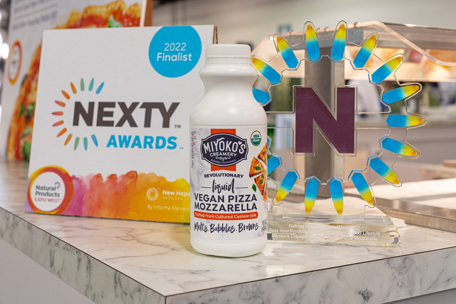 A bottle of Miyoko's Creamery liquid vegan pizza mozzarella sits beside a Nexty Awards 2022 finalist sign and trophy