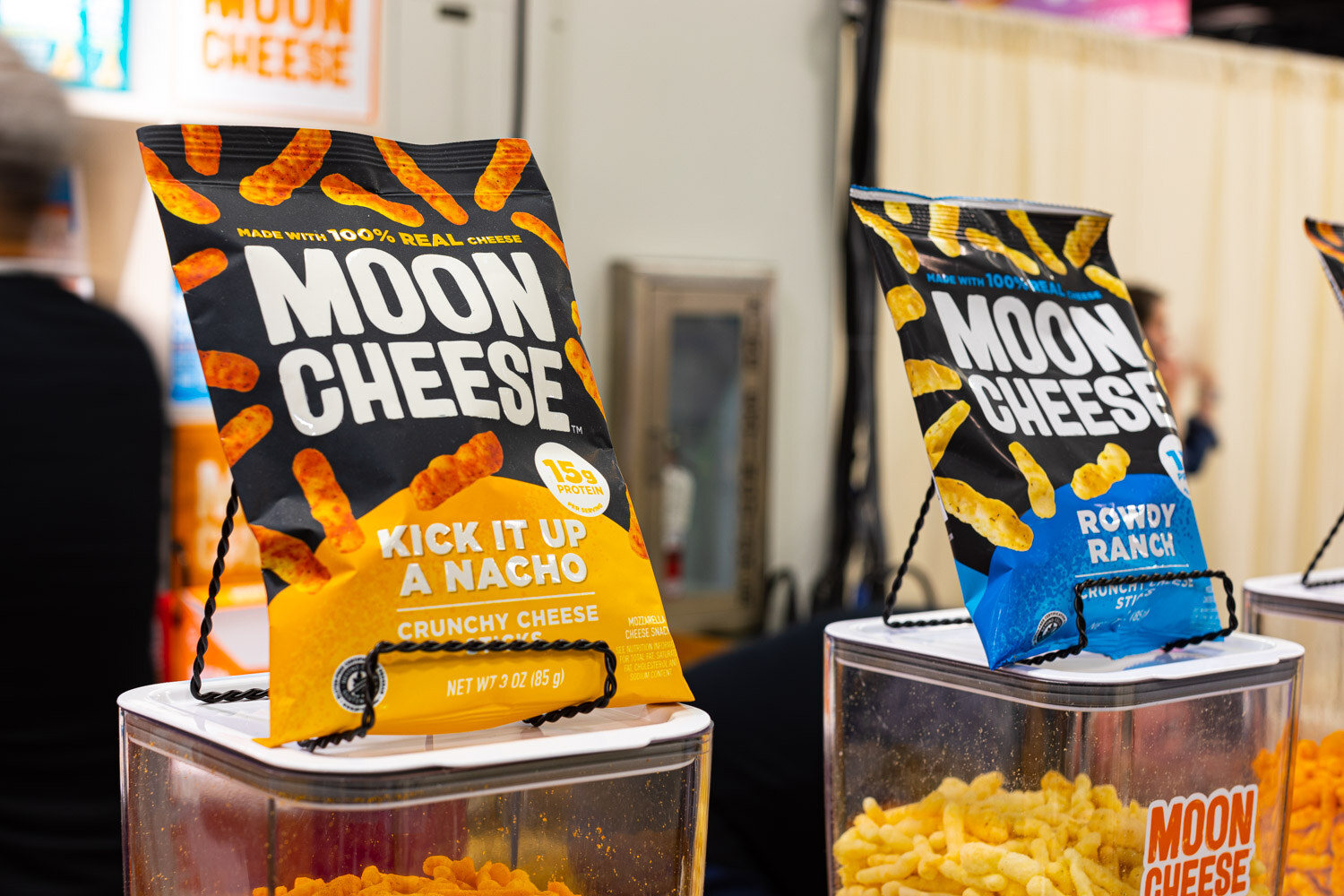 Bags of Moon Cheese Kick It Up a Nacho and Rowdy Ranch stand on top of containers of cheese snacks