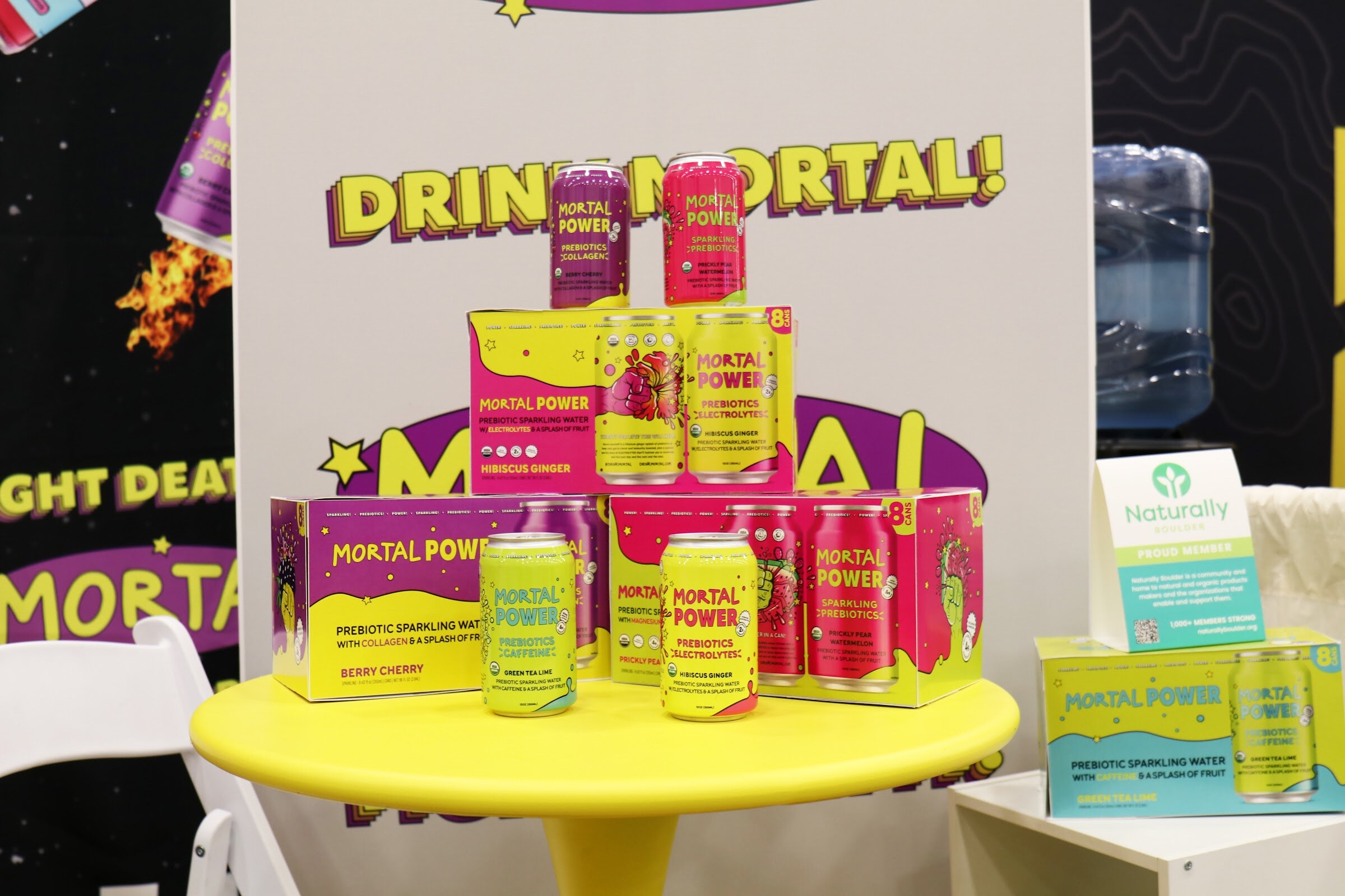 Boxes and cans of Mortal Power prebiotic sparkling water are stacked on a small yellow table in front of a Drink Mortal sign