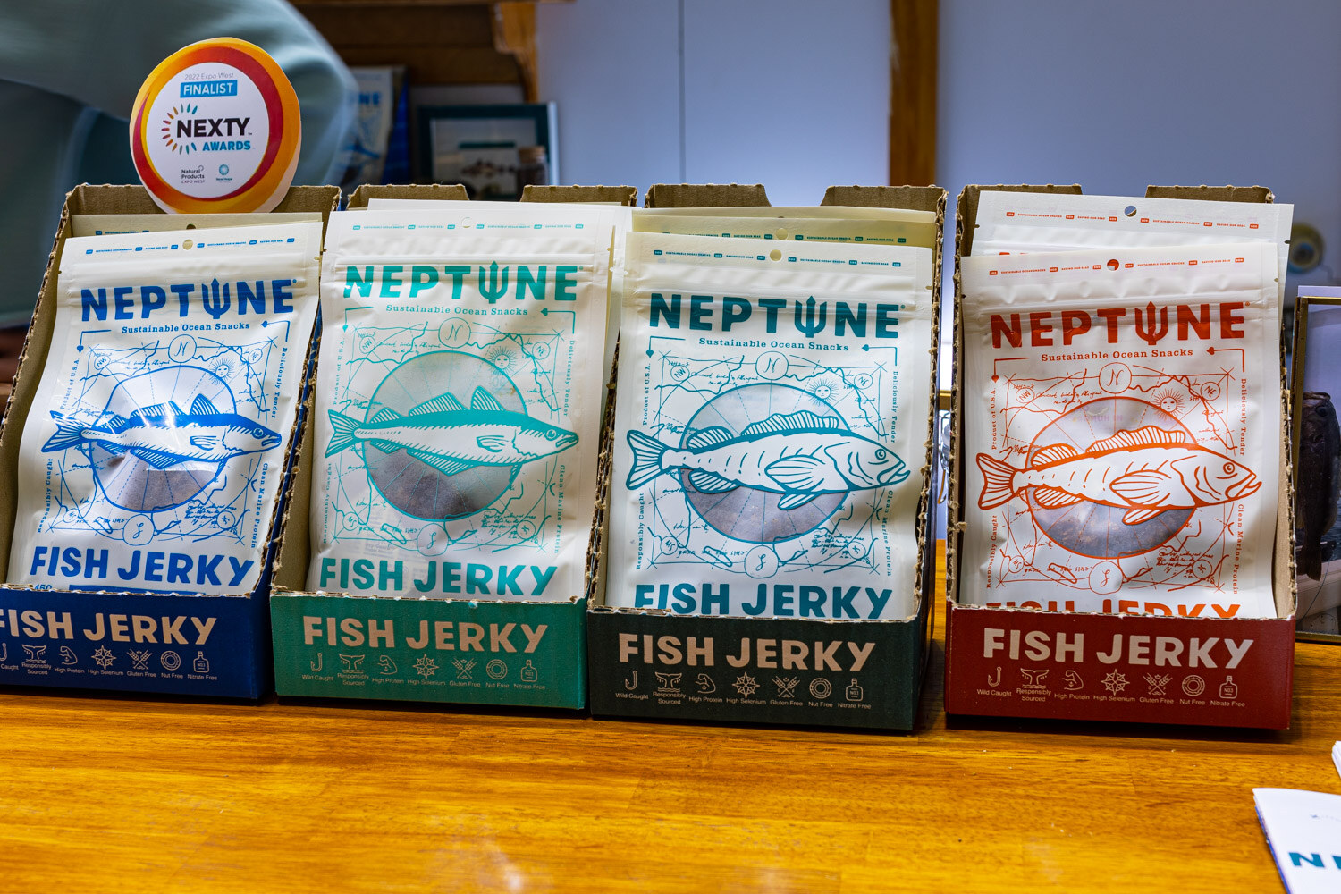 Four flavors of Neptune sustainable fish jerky stand in display boxes on a wooden counter under a Nexty finalist badge
