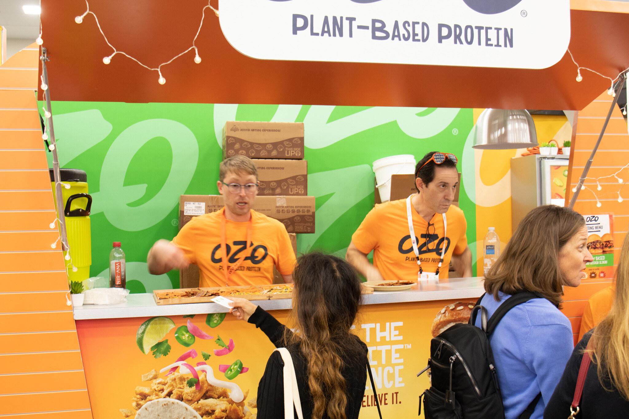 Two staff in orange Ozo shirts serve visitors at a colorful booth with a plant-based protein sign