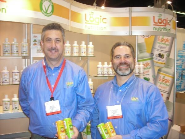Two men in blue Logic Nutrition shirts and show badges stand in front of shelves of Logic Nutrition bottles. One holds small green and orange cans.