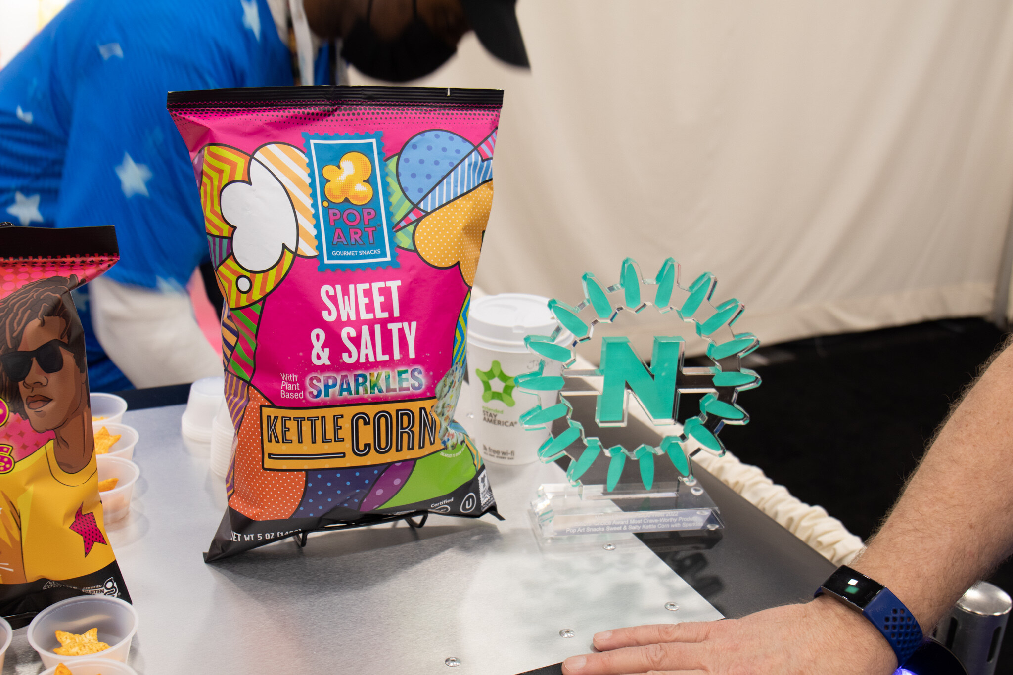 A bag of Pop Art Sweet and Salty Sparkles kettle corn stands next to a clear Nexty trophy as sample cups sit on the counter