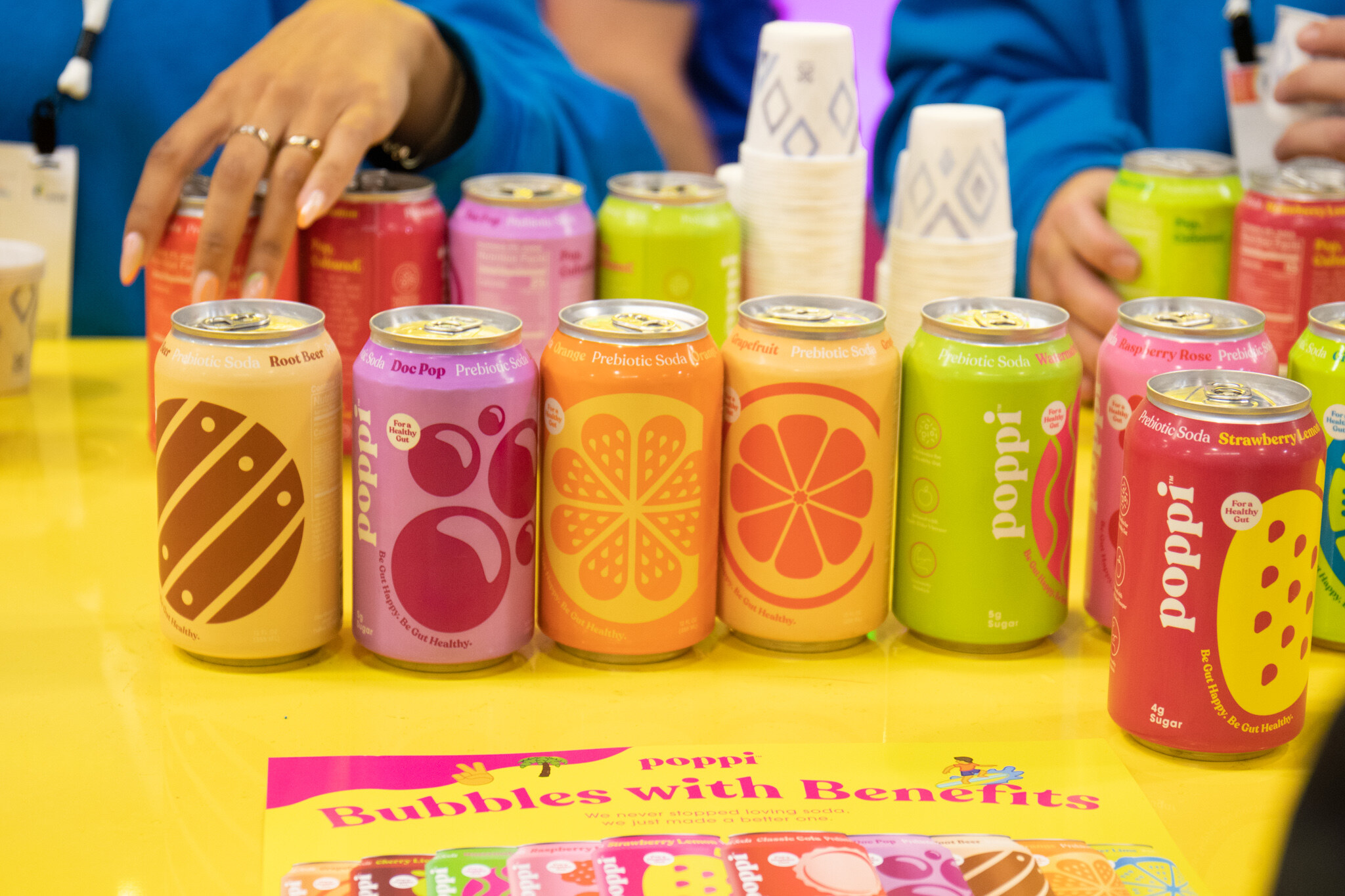Colorful cans of poppi prebiotic soda stand in a row on a yellow counter as staff hands arrange them