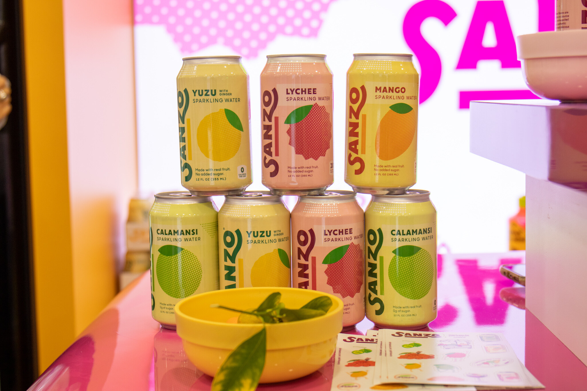 Cans of Sanzo yuzu, lychee, mango and calamansi sparkling water are stacked on a pink counter with a bowl of fruit