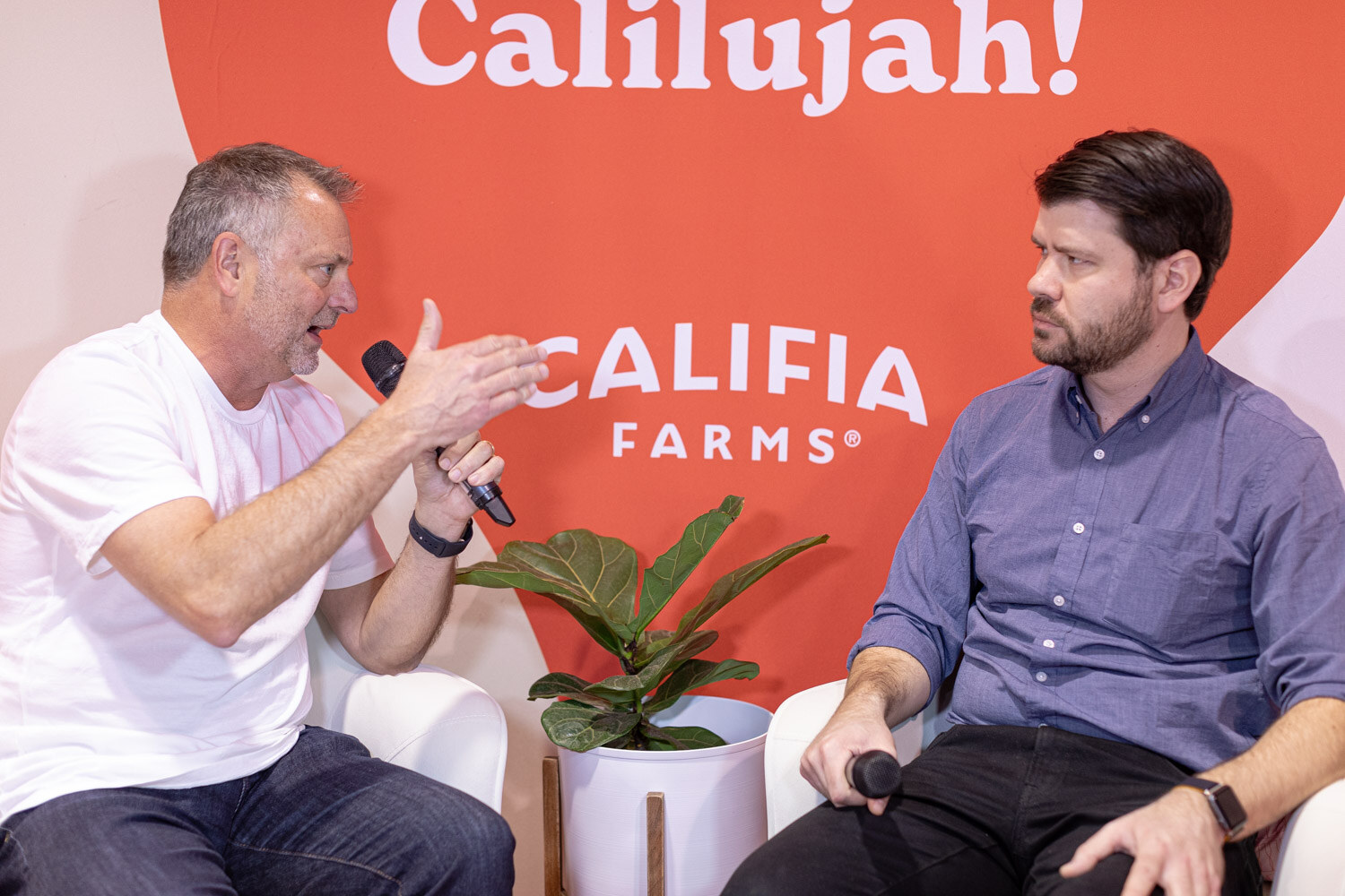 Two men sit in white chairs with microphones in front of a Califia Farms Calilujah backdrop during a video interview