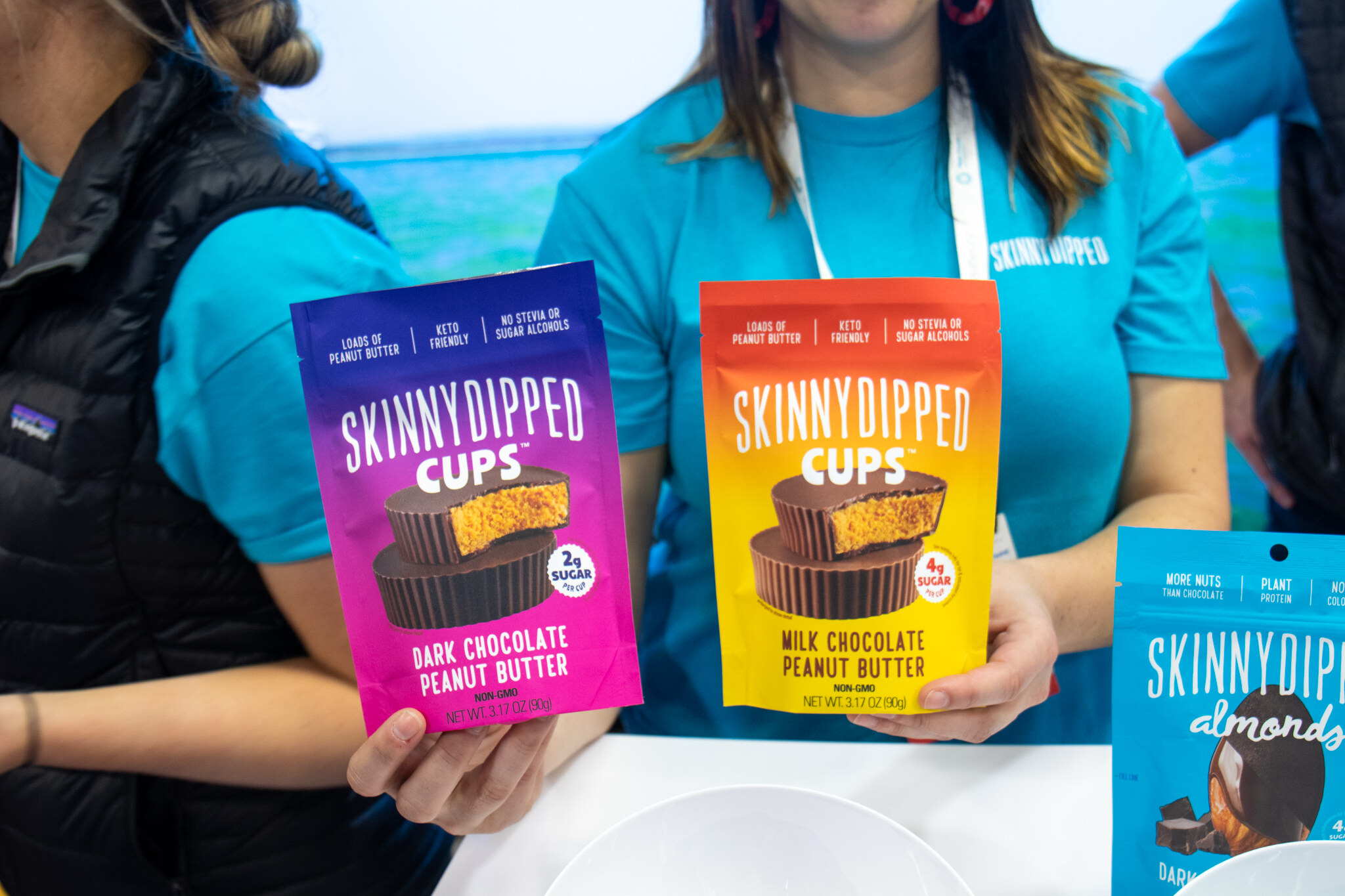 Two booth staff in teal shirts hold bags of SkinnyDipped dark chocolate and milk chocolate peanut butter cups