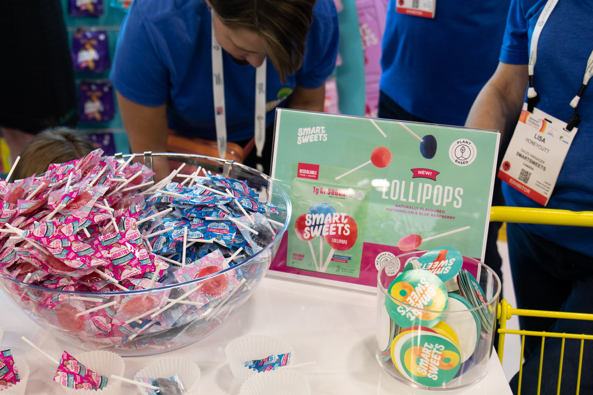 A glass bowl of wrapped lollipops sits in front of a Smart Sweets Lollipops sign as staff work in the background