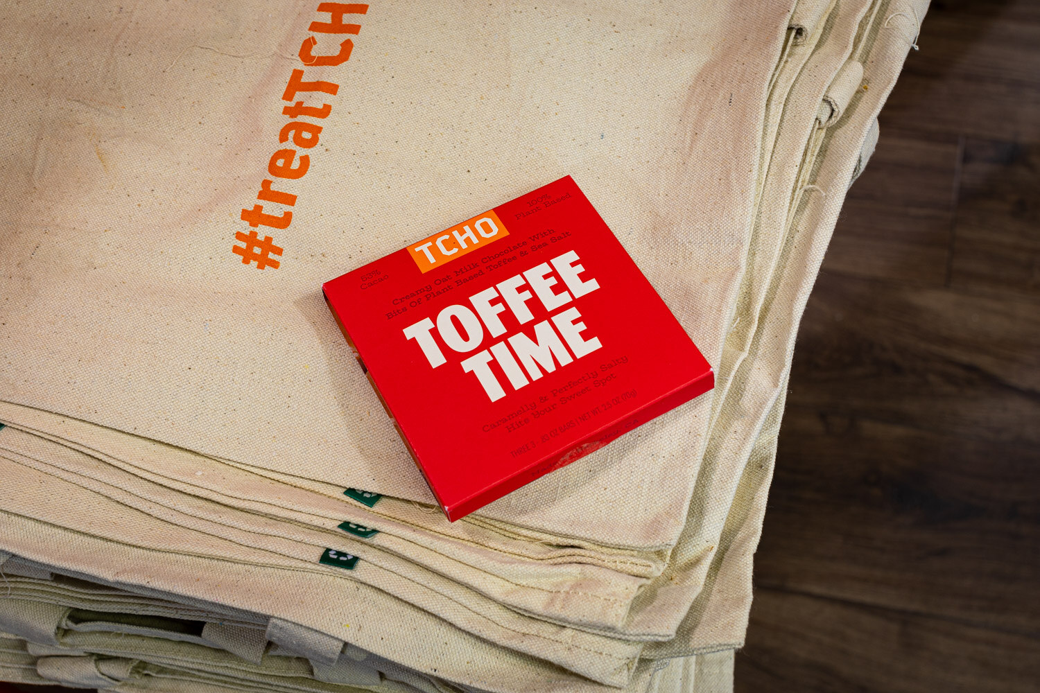 A red TCHO Toffee Time chocolate bar lies on a stack of canvas tote bags printed with #treatTCHO