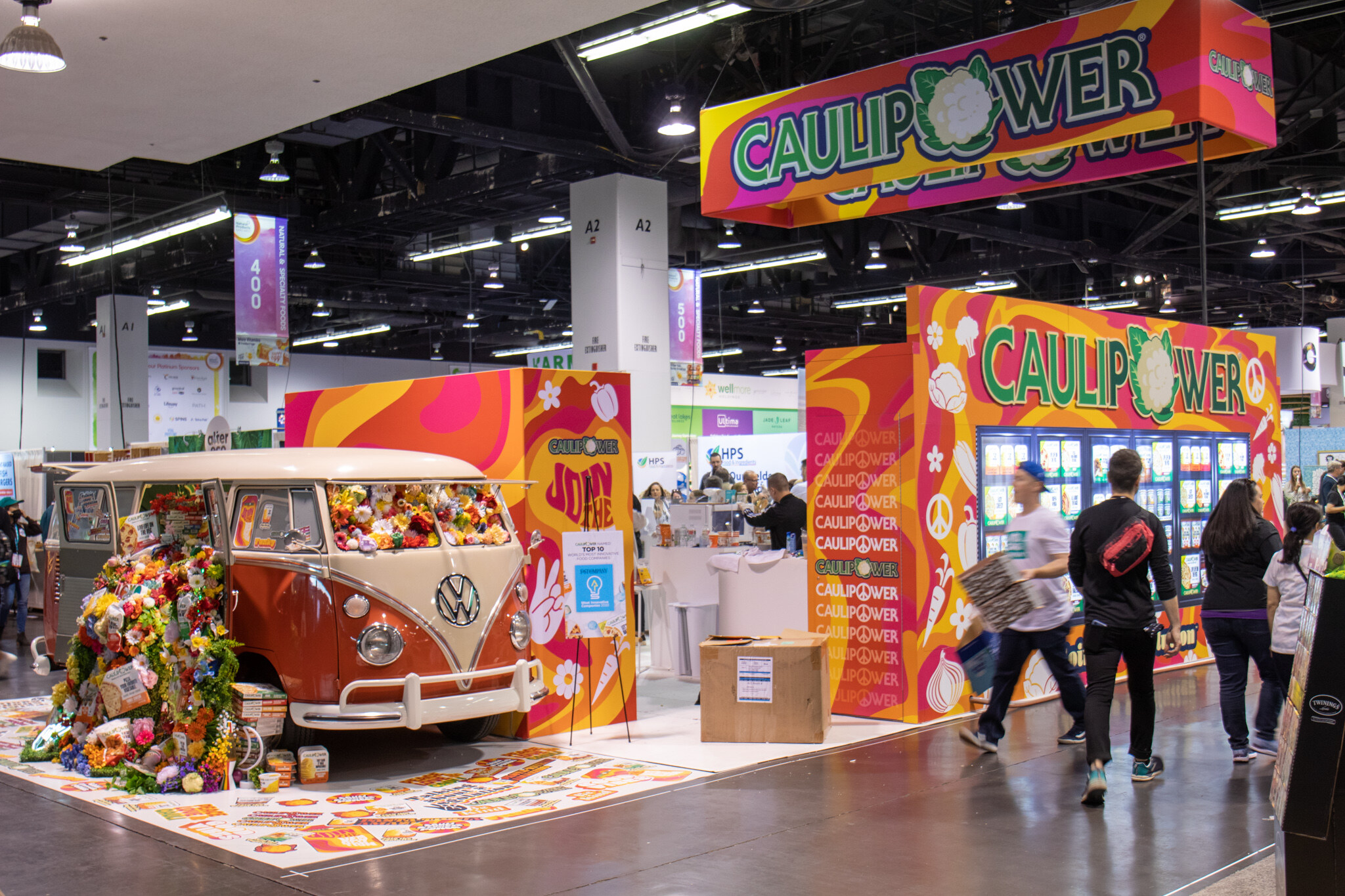 A wide view of the Caulipower booth with a vintage orange Volkswagen van covered in flowers beside a colorful branded wall and freezer