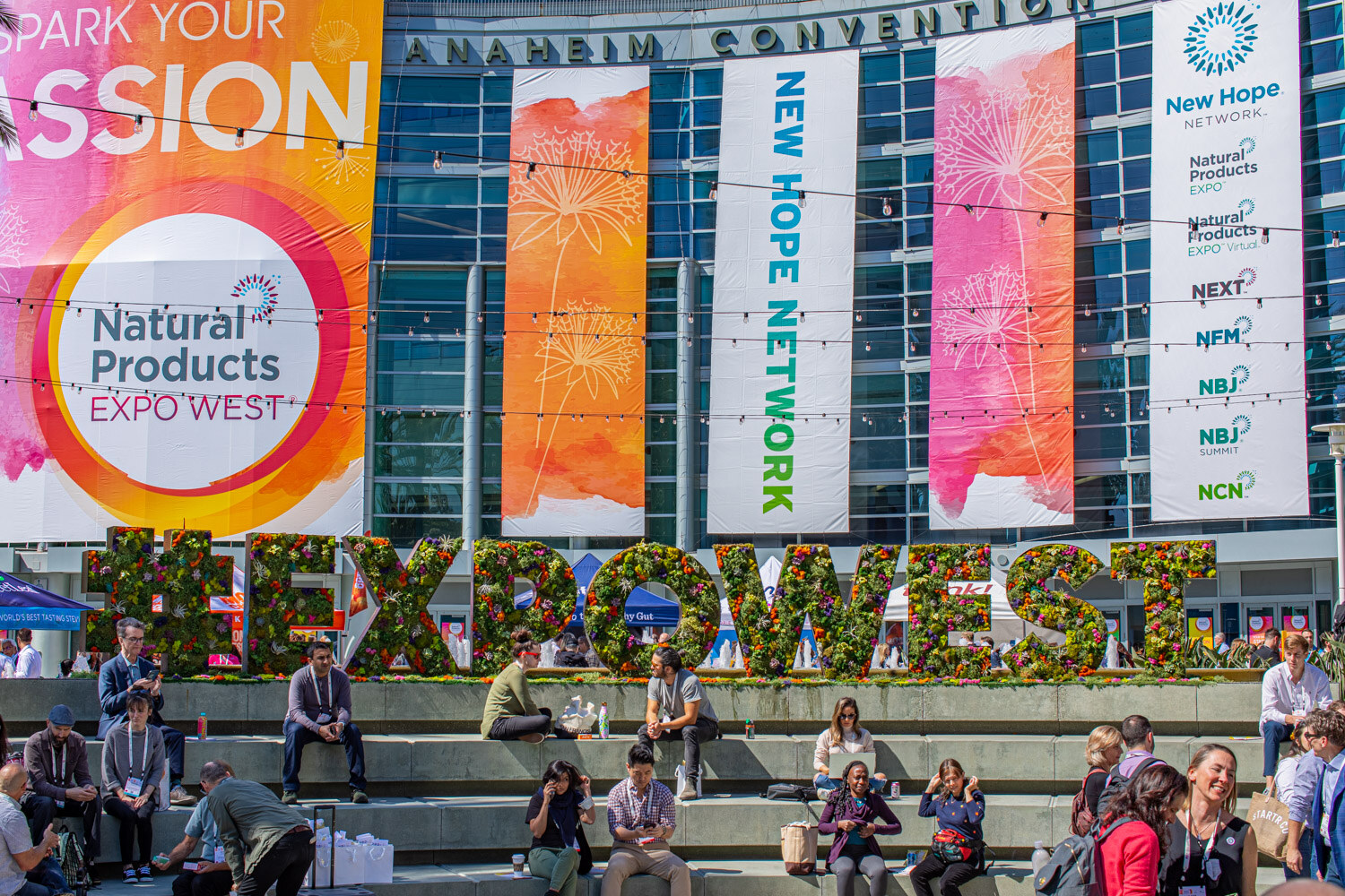 People sit on steps in front of a floral #EXPOWEST letter sign beneath Natural Products Expo West and New Hope Network banners