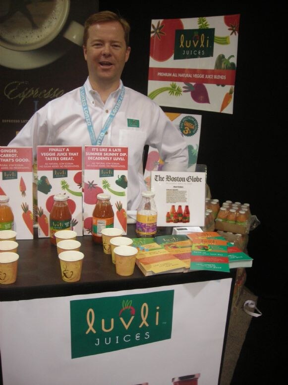 A man in a white shirt and lanyard stands behind a Luvli Juices table with veggie juice bottles, paper sample cups, brochures and a Boston Globe article display.