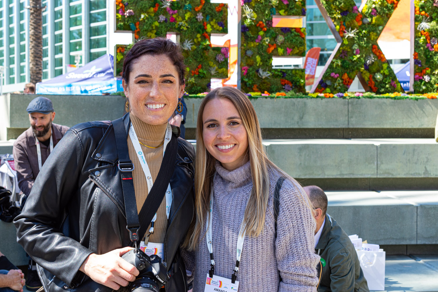 Two smiling women with lanyards, one holding a camera, pose outdoors in front of a floral EXPOWEST letter sign