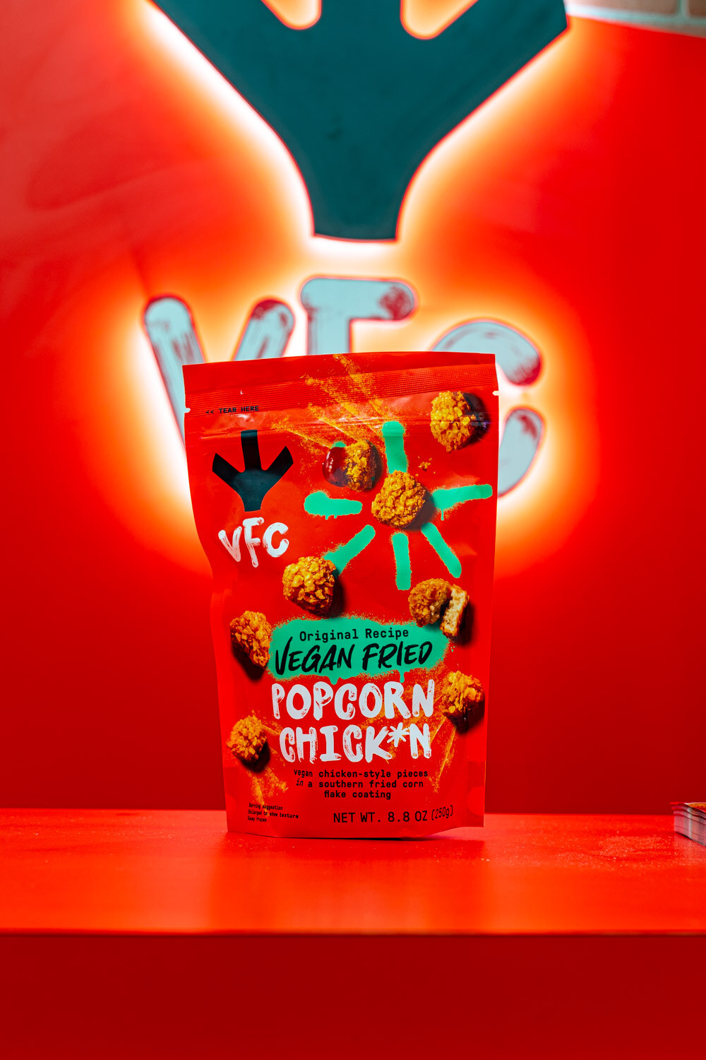 A bag of VFC Vegan Fried Popcorn Chick'n stands in front of a glowing red VFC sign