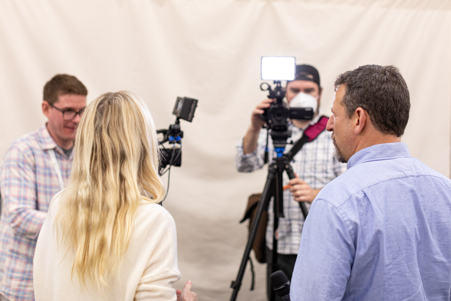 A camera operator films two men and a blonde woman in front of a cream backdrop as an interview is set up