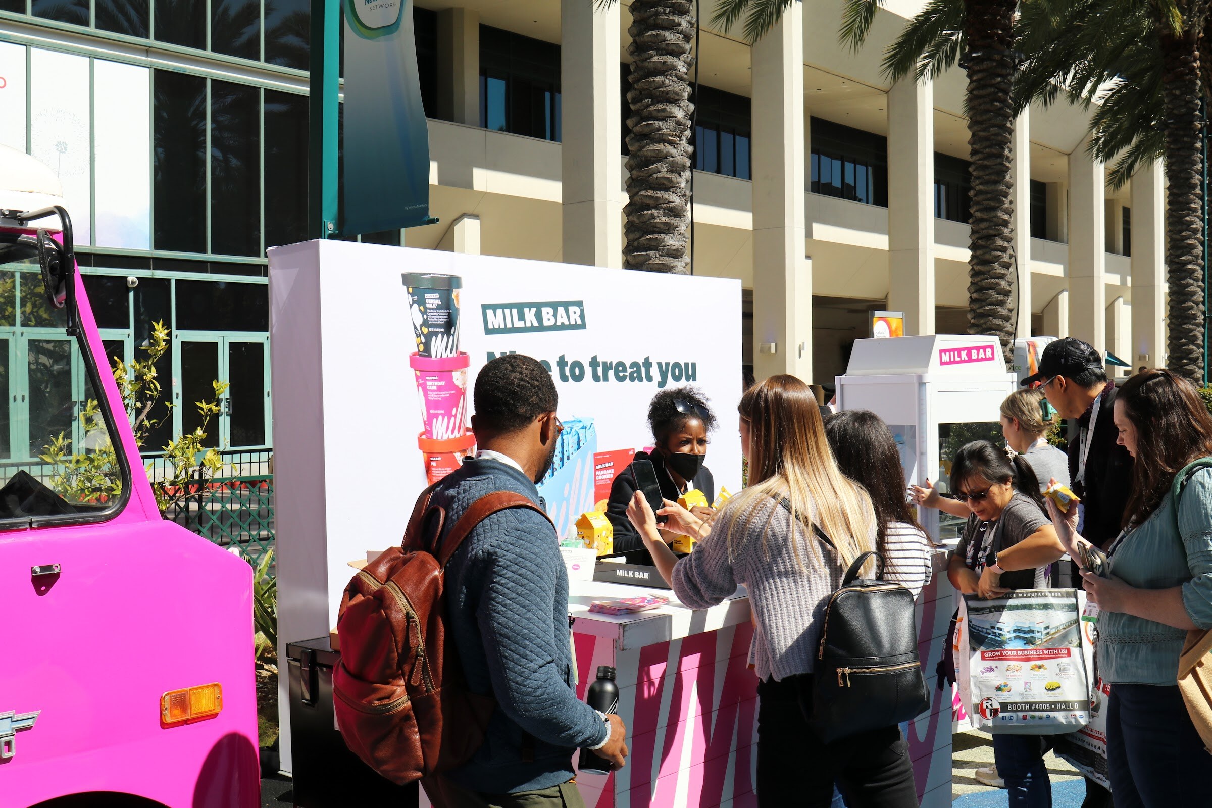 Attendees crowd around a pink Milk Bar truck with a white serving counter on the outdoor plaza