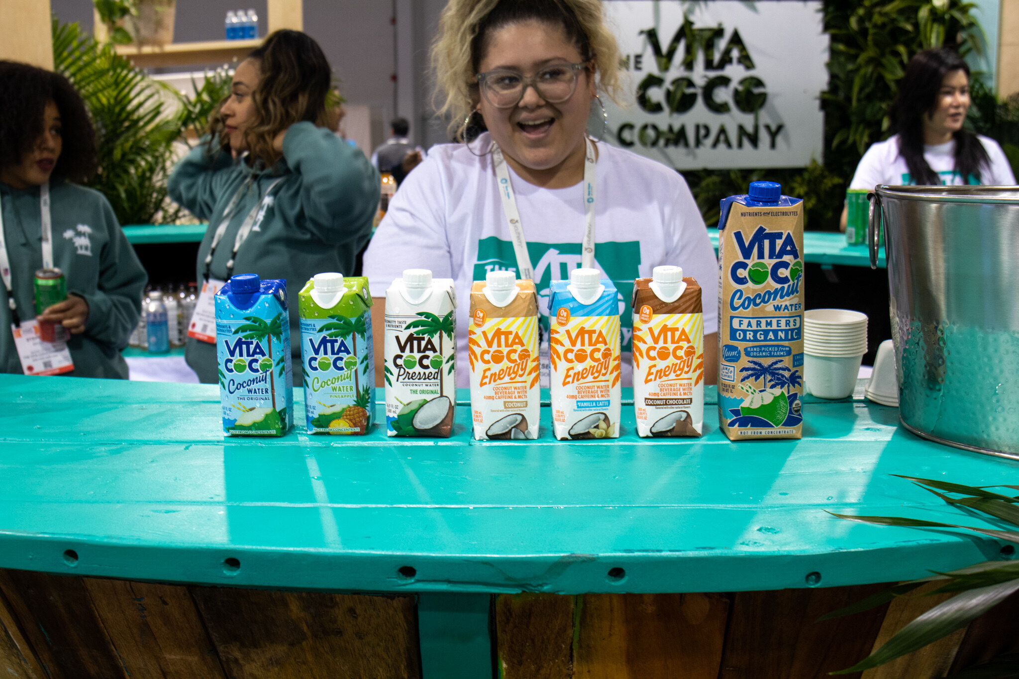A smiling woman stands behind a teal counter lined with Vita Coco coconut water and energy cartons