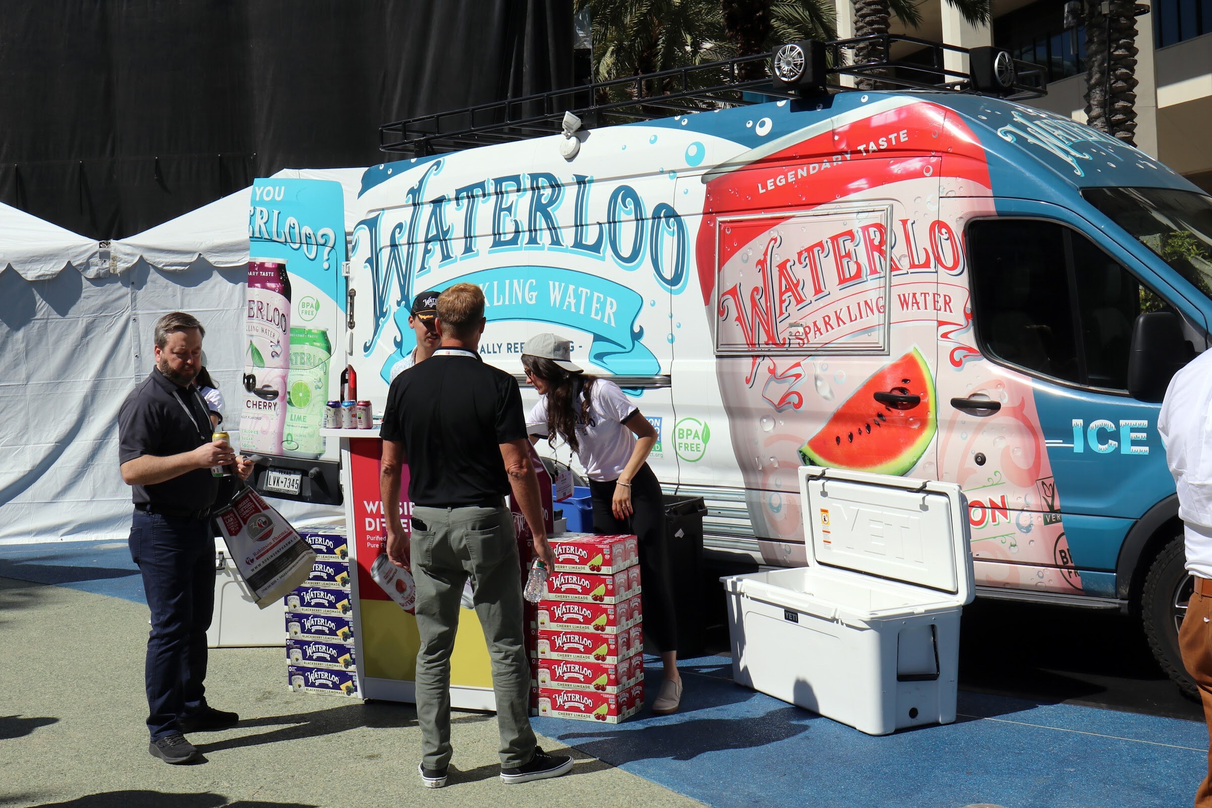 Staff and visitors stand beside a Waterloo Sparkling Water van with stacked cases and a cooler on a blue paved plaza