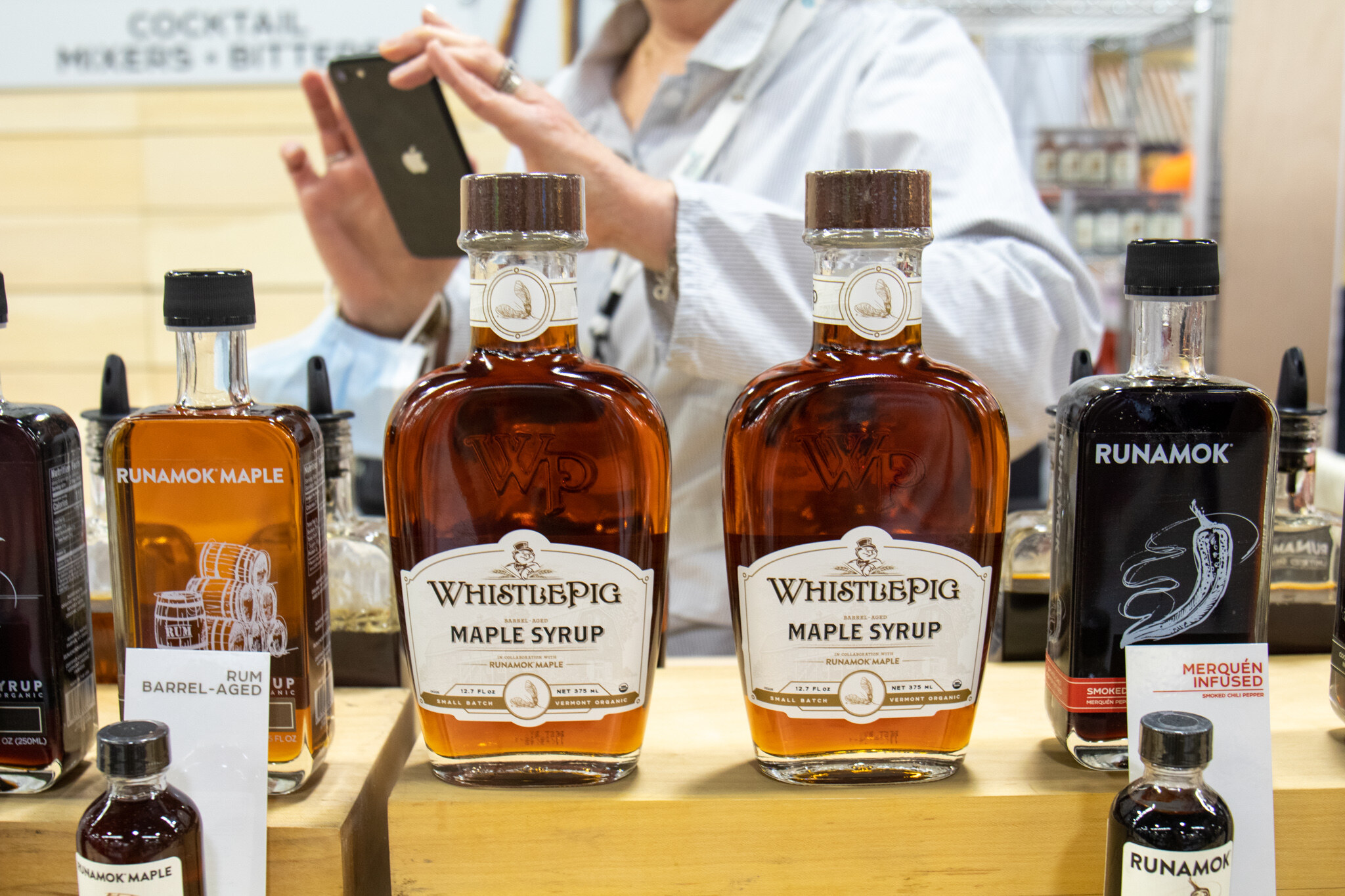 Bottles of WhistlePig maple syrup and Runamok maple and infused syrups stand on a wooden counter while someone photographs them with a phone