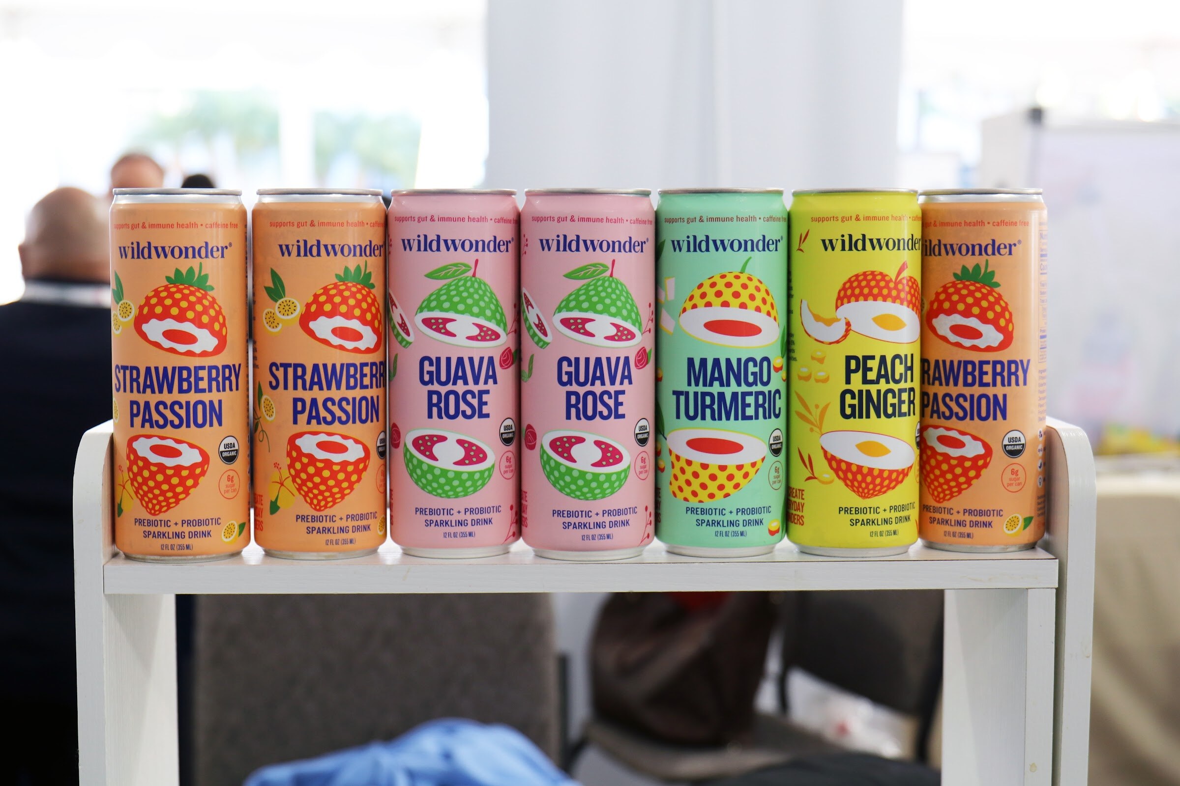 A row of Wildwonder prebiotic sparkling drink cans in strawberry passion, guava rose and mango turmeric flavors stands on a small white shelf