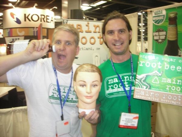 Two men in Maine Root shirts pose together. One holds a bottle to his ear and mimics shouting, the other holds a mannequin head. A Maine Root root beer sign is at right.
