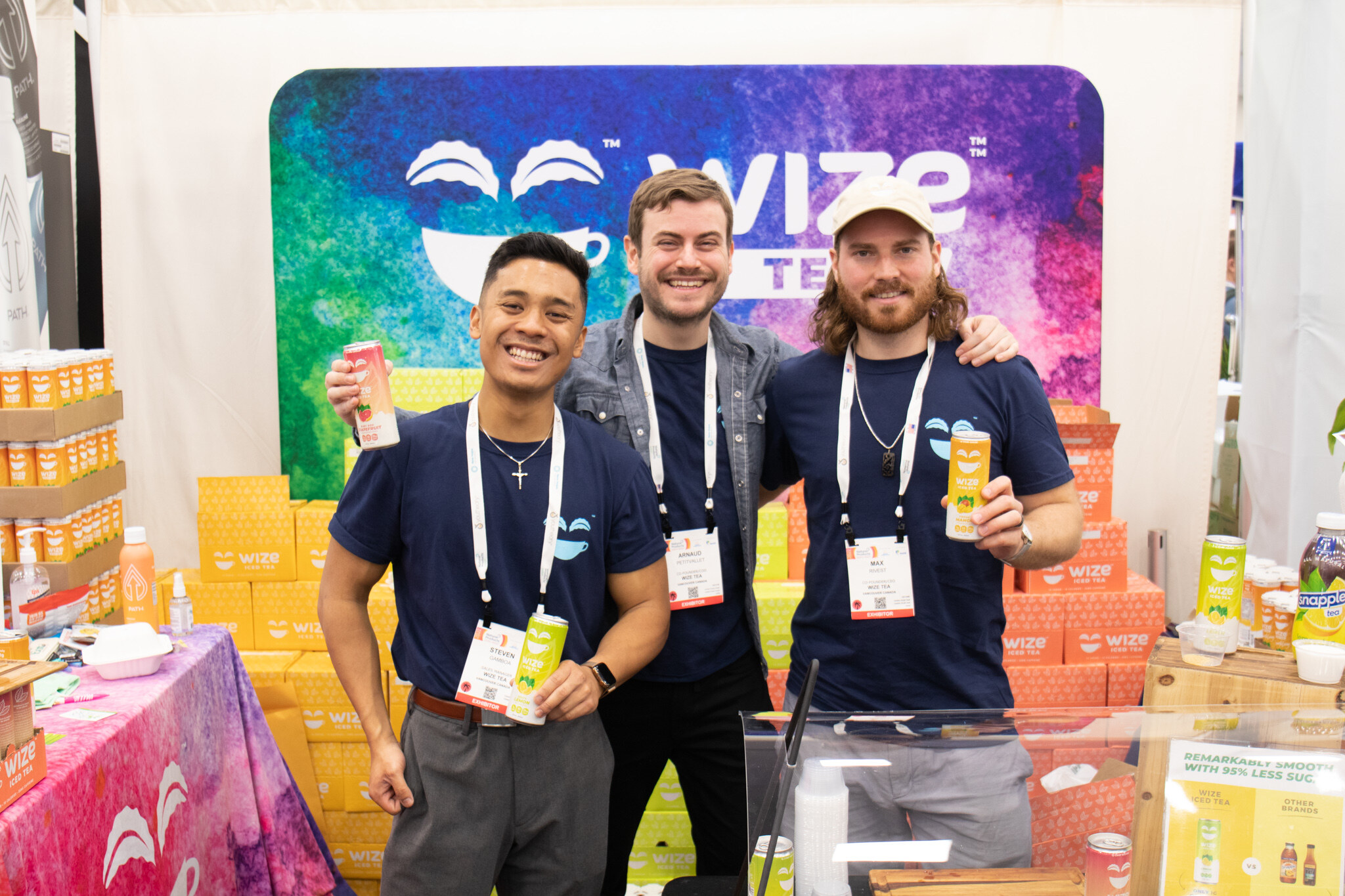 Three smiling booth staff with lanyards hold up cans in front of a colorful Wize backdrop and stacked cases