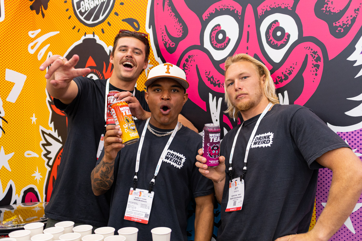 Three team members in matching dark shirts and lanyards pose in front of a colorful illustrated booth wall holding drinks