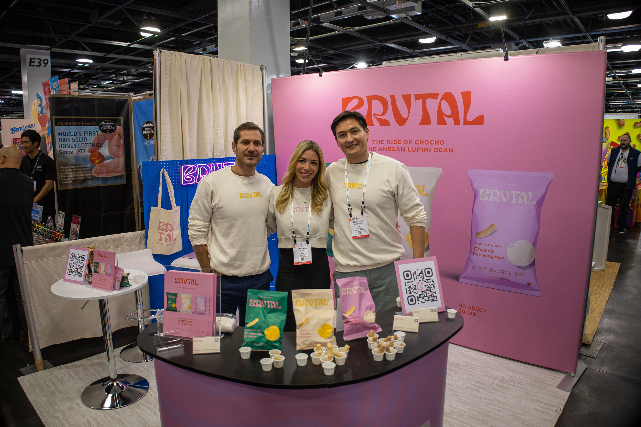 Three people in matching cream shirts stand in a pink Brutal booth with bags of snacks on the counter