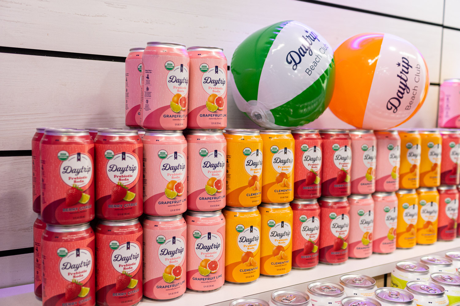 Stacked Daytrip prebiotic soda cans in red, pink and orange next to beach balls