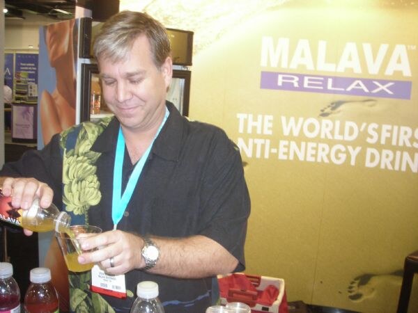 A smiling man in a dark patterned shirt and teal lanyard pours a yellow drink from a bottle into a cup in front of a Malava Relax sign.