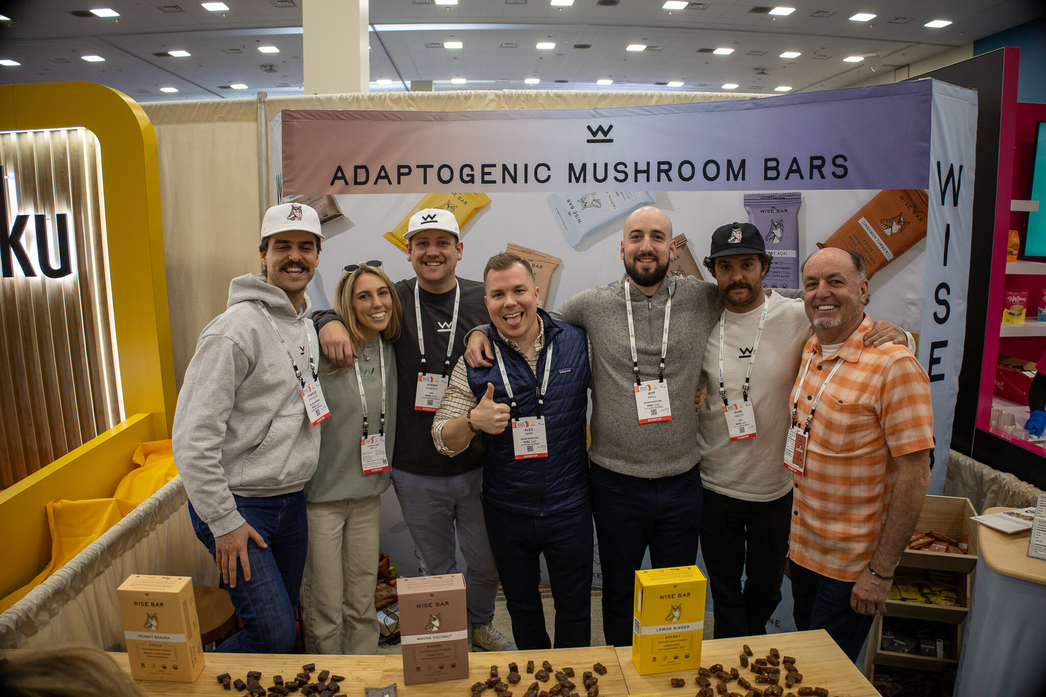 A group of eight people stand together smiling in a booth with a sign reading Adaptogenic Mushroom Bars