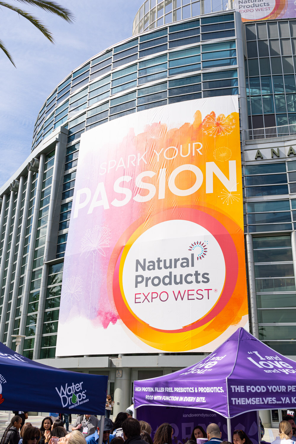 A large Natural Products Expo West banner on the glass facade of the Anaheim Convention Center with an attendee crowd below