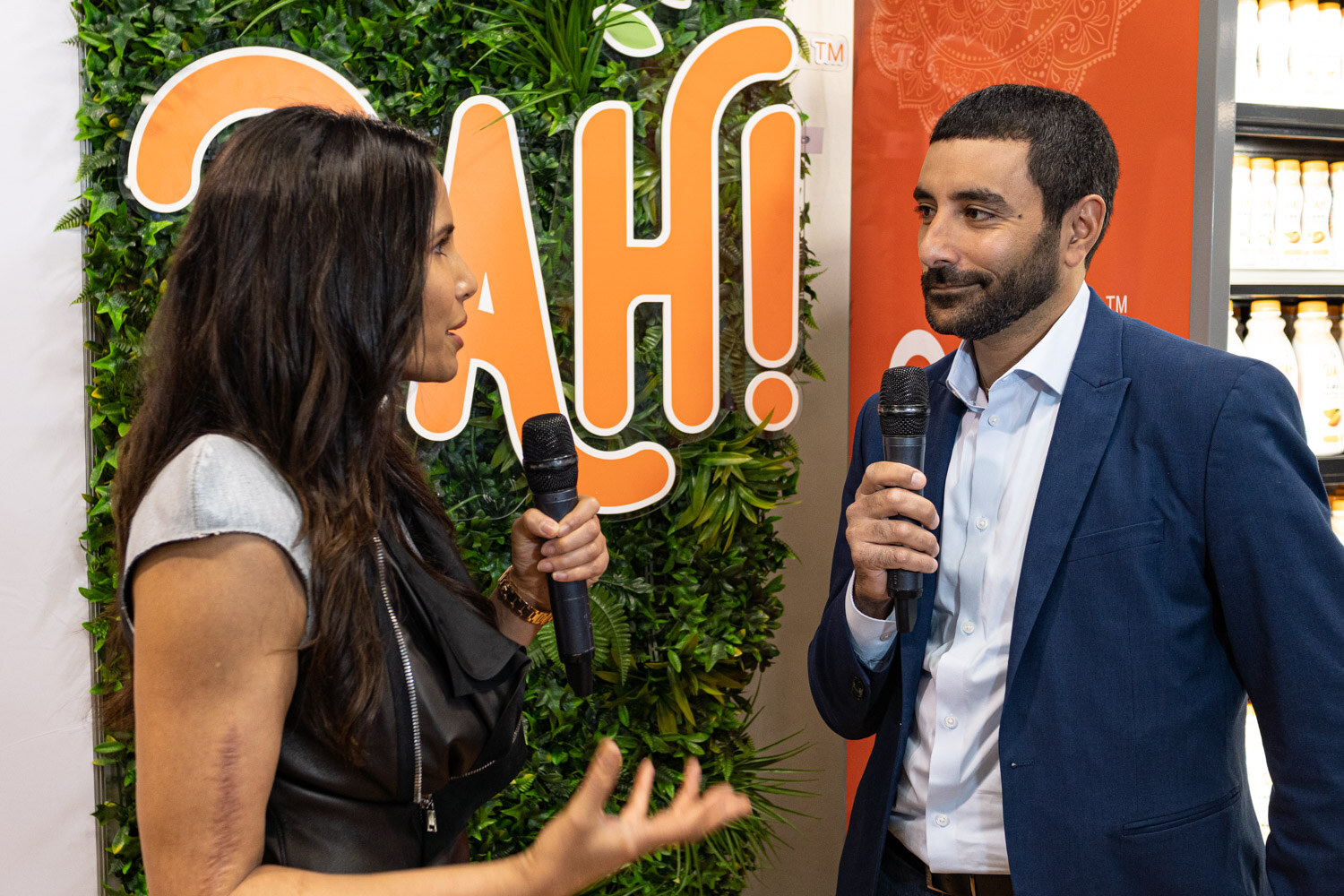 Two people holding microphones talk in front of a green plant wall with an orange Dahi sign