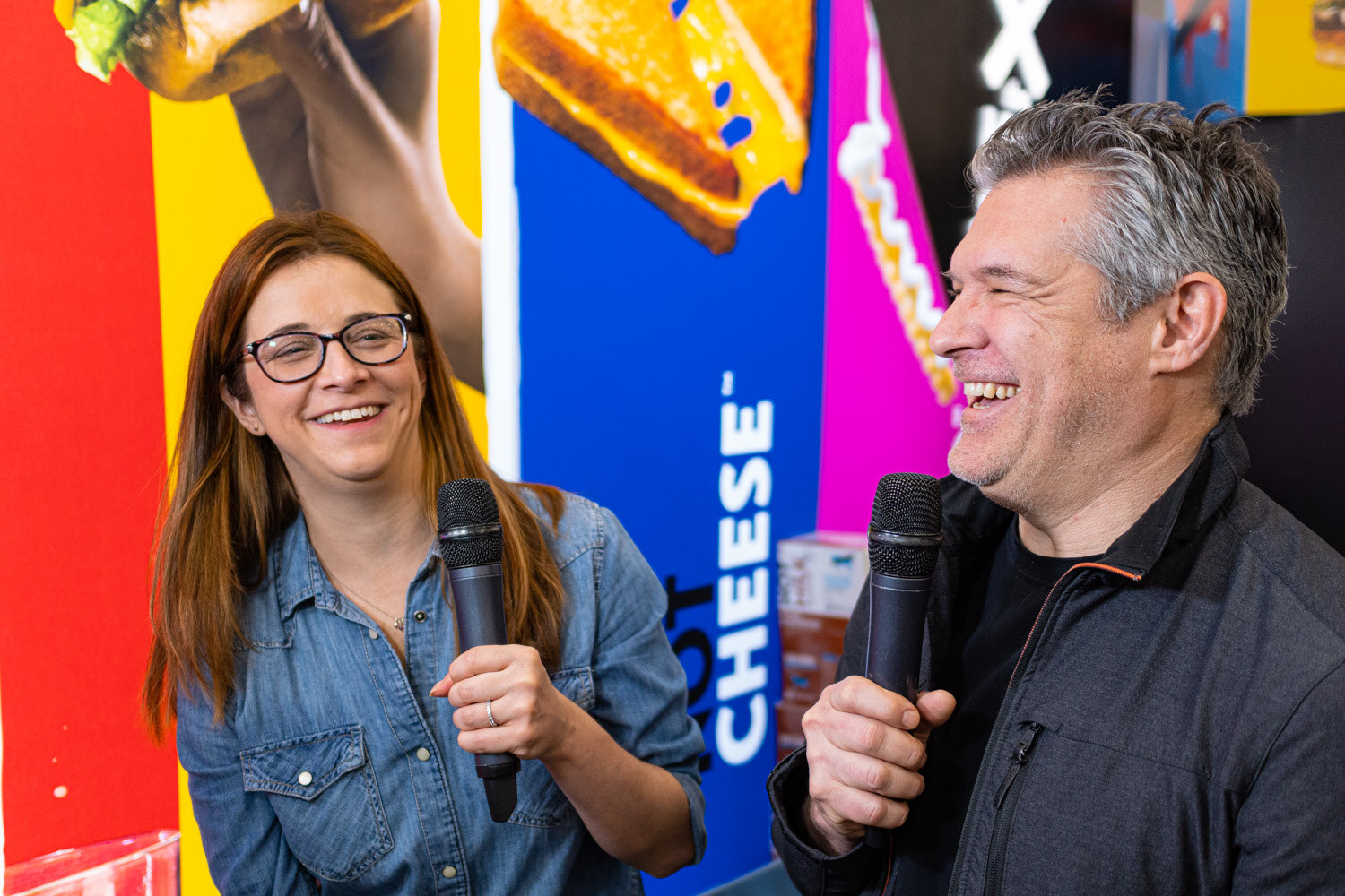 Two people hold microphones and laugh during an interview in front of a booth wall with a cheese graphic