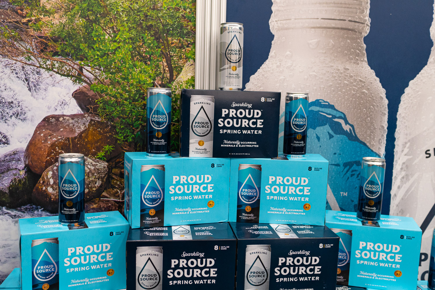 Stacked Proud Source sparkling water cartons and cans in front of a mountain stream photo