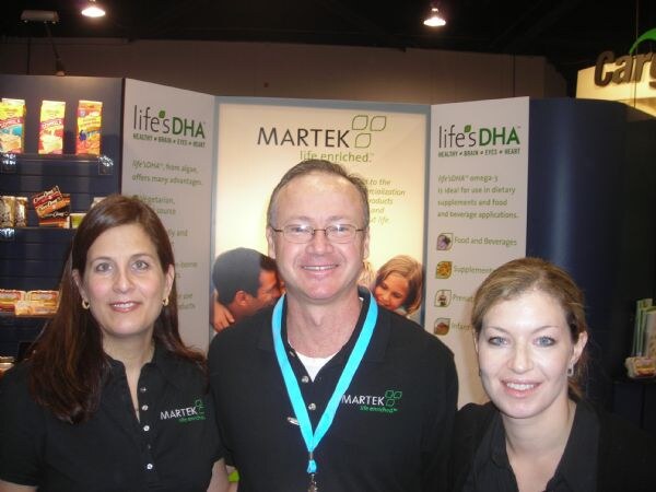 Two women and a man in Martek shirts stand together in front of Martek and life's DHA display panels. The man wears glasses and a teal lanyard.