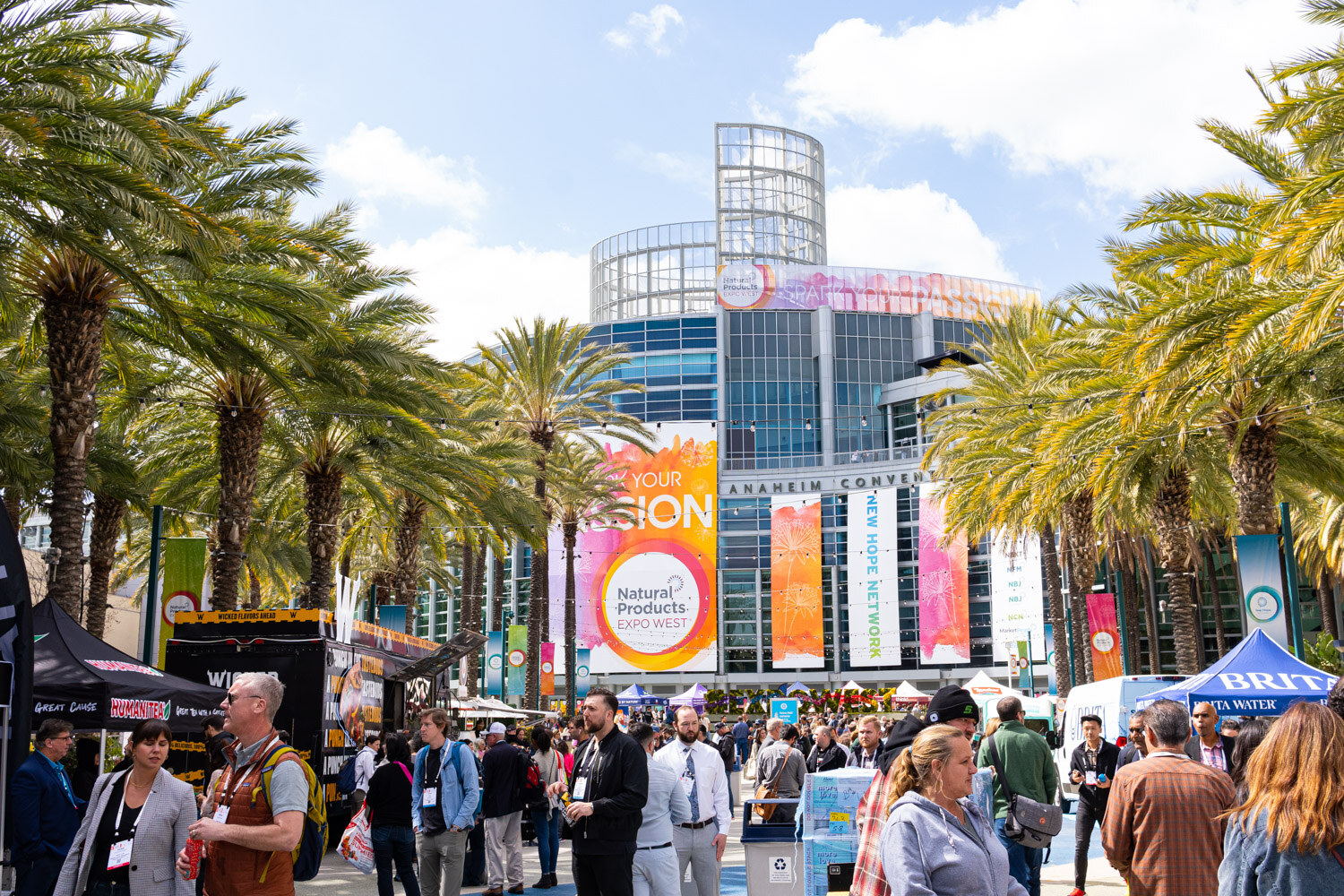 Crowds of badge-wearing attendees walk through palm trees toward the Anaheim Convention Center with show banners overhead