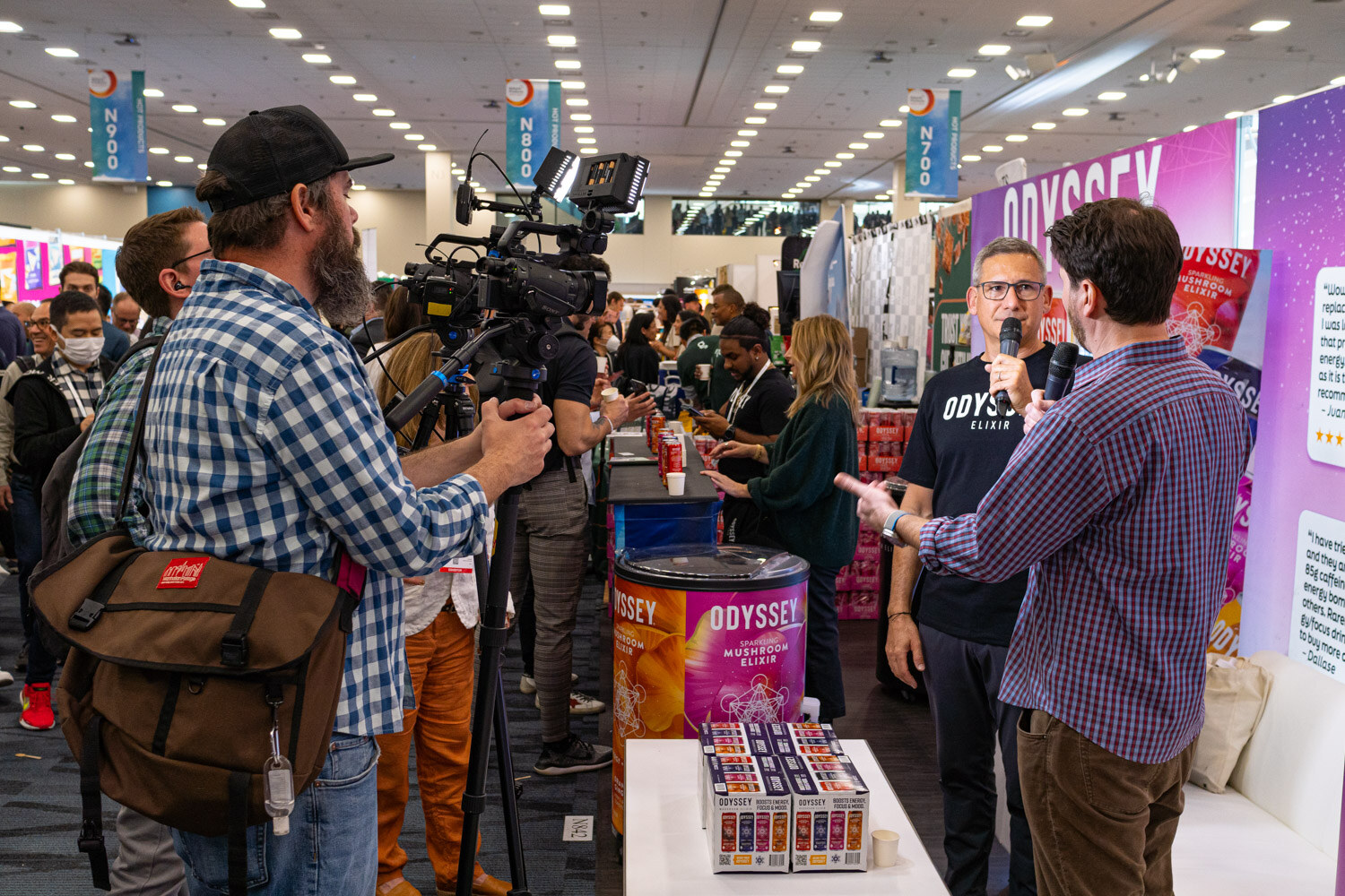 A camera operator films a person being interviewed at a booth while onlookers watch