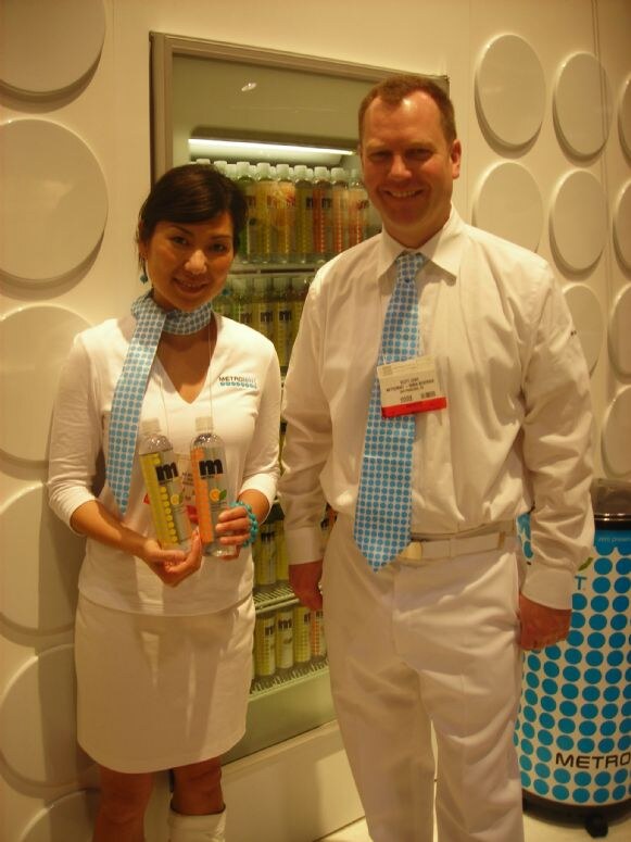 A woman and a man in white clothing and matching blue polka-dot ties stand by a lit cooler of drinks. The woman holds two Metromint bottles.