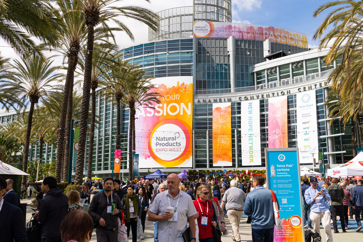 Attendees gather on the plaza in front of the Anaheim Convention Center beneath large Natural Products Expo West banners reading Spark Your Passion