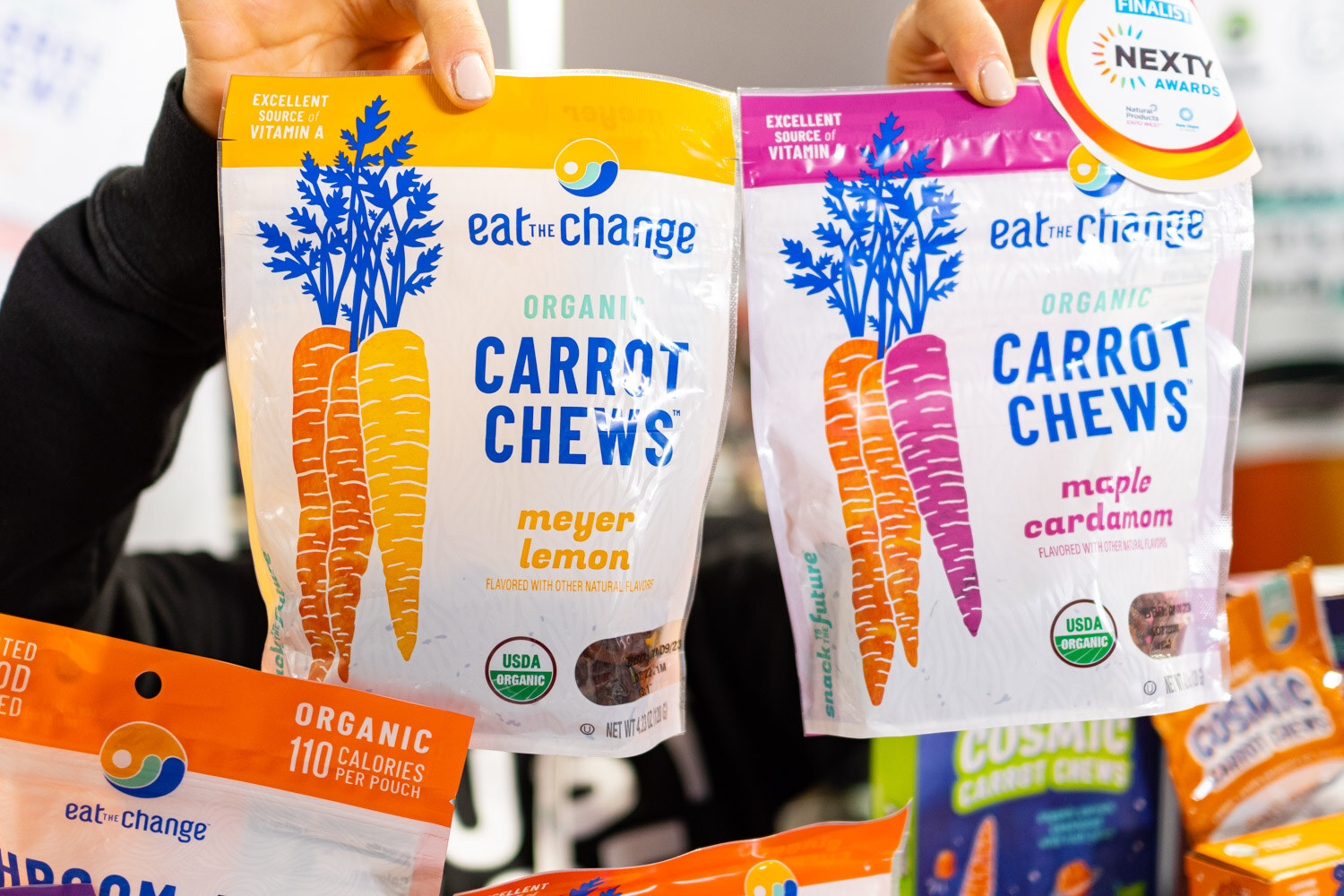 Two bags of eat. change organic carrot chews held up in front of a booth