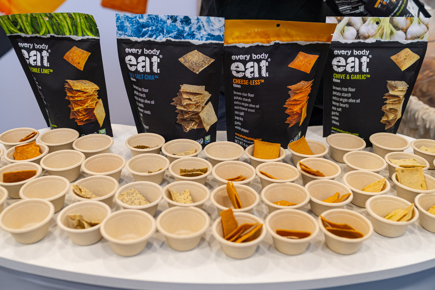 Bags of Every Body Eat crackers behind a table full of small sample cups