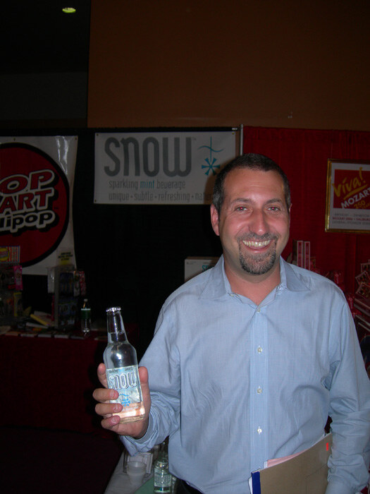 A smiling man with a goatee in a light blue shirt holds a bottle of Snow and a folder in front of a Snow sparkling mint beverage sign.