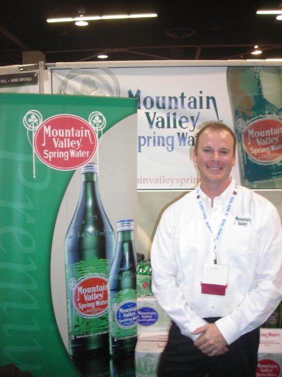 A smiling man in a white Mountain Valley shirt and lanyard stands beside banners showing Mountain Valley Spring Water glass bottles.