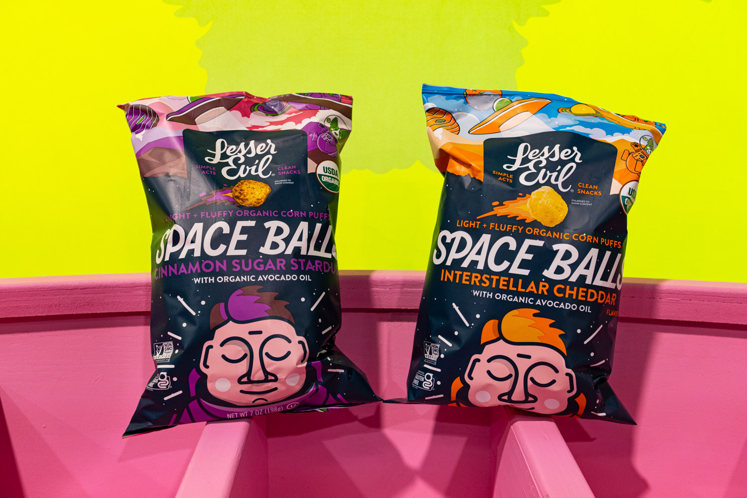 Two bags of Lesser Evil Space Balls on a pink display against a bright yellow background