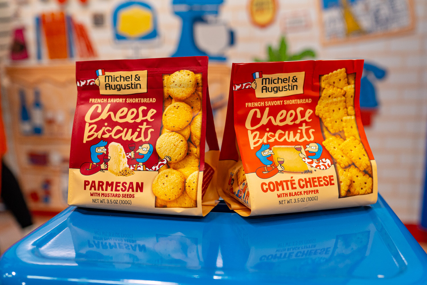 Two bags of Michel et Augustin Parmesan and Comte cheese biscuits on a blue display