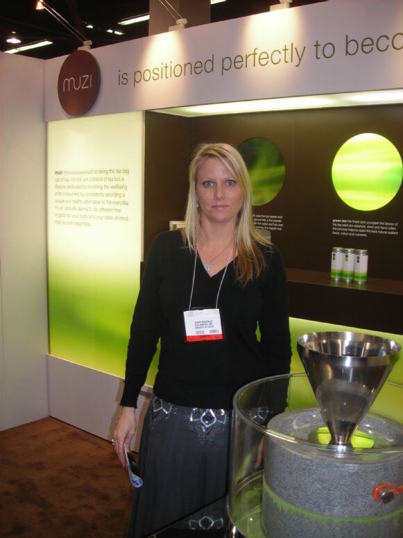 A blonde woman in a black top and show badge stands in the muzi booth, which has green lit panels, small tea tins and a stone grinder with a steel funnel in front.